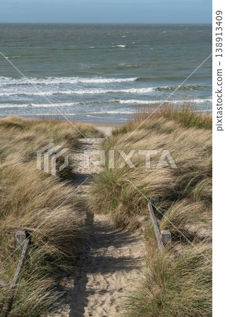 Sandy path through coastal dunes leading to North Sea waves with grass and shoreline under clear sky. Travel news, tourism coverage, seaside destination, coastal Sandy path through coastal dunes leading to North Sea waves with grass and shoreline under clear sky. Travel news, tourism coverage, seaside destination, coastal 138913409