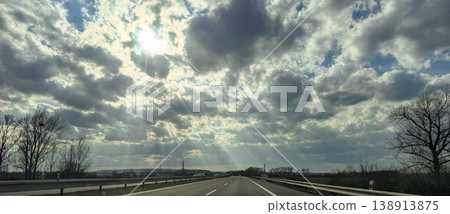 Highway road beneath cloudy sky with sunrays over rural landscape for travel transport and weather concept Highway road beneath cloudy sky with sunrays over rural landscape for travel transport and weather concept 138913875