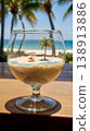 Tiny tropical island with palm and umbrella in cocktail glass. wooden table with real ocean and palm trees in the background. 138913886