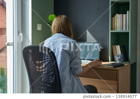 Woman sitting at desk and using laptop in home office near window Woman sitting at desk and using laptop in home office near window 138914077