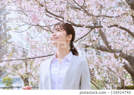 A young woman in a suit smiles under cherry blossoms in full bloom - an image of spring and new beginnings - the season for new employee induction ceremonies. 138914740
