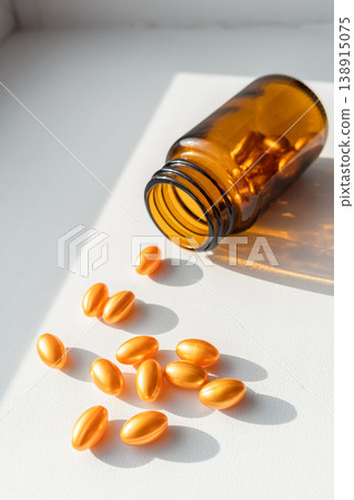 Amber bottle spilling golden softgel capsules on white background, representing medicine, vitamins, and skincare. Bottle spilling golden pills for health and beauty 138915075