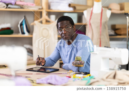 Male tailor draws sketch of future clothing on an electronic tablet. Working with modern gadgets in sewing workshop Male tailor draws sketch of future clothing on an electronic tablet. Working with modern gadgets in sewing workshop 138915750