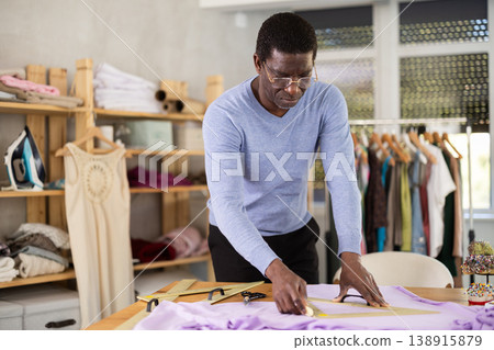 Male tailor draws outline of future dress on fabric with chalk in sewing workshop 138915879