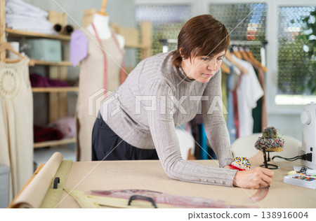 Female dressmaker draws sketch of future clothing on paper using patterns in sewing workshop Female dressmaker draws sketch of future clothing on paper using patterns in sewing workshop 138916004