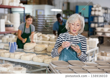 Portrait of elderly woman in apron cutting burrs off freshly made earthenware cups and plates with knife Portrait of elderly woman in apron cutting burrs off freshly made earthenware cups and plates with knife 138916010