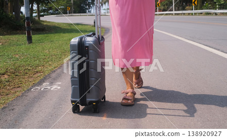 Young woman in pink dress walking along a road pulling a grey suitcase with lush tropical trees in the background. Vacation travel concept 138920927