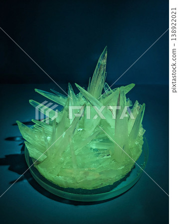 A close up view of sharp, translucent green crystals forming in a glass petri dish under blue light, showcasing natural geometric growth patterns 138922021
