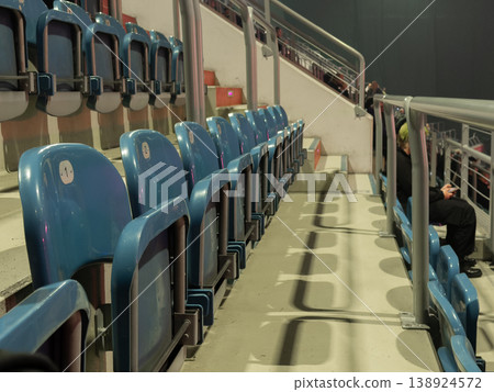 Modern blue stadium seats stand mostly empty with a solitary figure quietly using a phone, representing contemplation and anticipation at a public event Modern blue stadium seats stand mostly empty with a solitary figure quietly using a phone, representing contemplation and anticipation at a public event 138924572