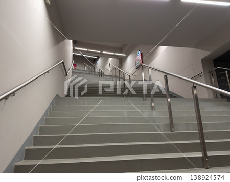Long, wide, grey concrete staircase with bright overhead illumination and sleek silver metal handrails emphasizes modern public architectural design and upward progression Long, wide, grey concrete staircase with bright overhead illumination and sleek silver metal handrails emphasizes modern public architectural design and upward progression 138924574