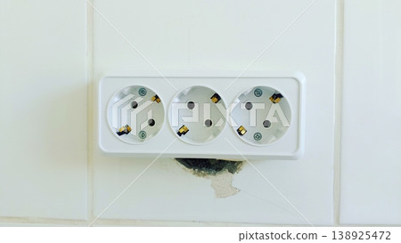 A front-view minimalist shot of a white triple electrical wall socket. Mounted on a clean white tiled wall with visible installation detail. A front-view minimalist shot of a white triple electrical wall socket. Mounted on a clean white tiled wall with visible installation detail. 138925472