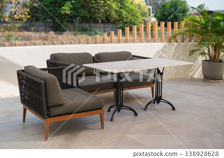 Modern outdoor furniture arrangement featuring stylish chairs and a sleek table on a sunny patio with greenery in the background Modern outdoor furniture arrangement featuring stylish chairs and a sleek table on a sunny patio with greenery in the background 138928628