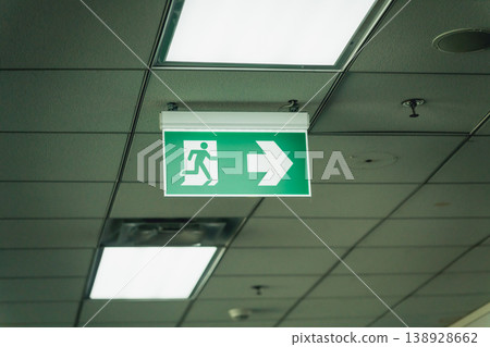 Green emergency exit sign hanging from ceiling indicating direction with illuminated interior lights and visible tiles for safety awareness and navigation Green emergency exit sign hanging from ceiling indicating direction with illuminated interior lights and visible tiles for safety awareness and navigation 138928662