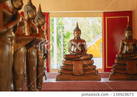 Serene Buddha Statues in a Tranquil Setting with Natural Light Filtering Through Vibrant Red Windows and Lush Greenery in Background Serene Buddha Statues in a Tranquil Setting with Natural Light Filtering Through Vibrant Red Windows and Lush Greenery in Background 138928673