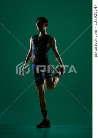 Male athlete stretching leg in standing position on dark green studio background. Male athlete stretching leg in standing position on dark green studio background. 138929147