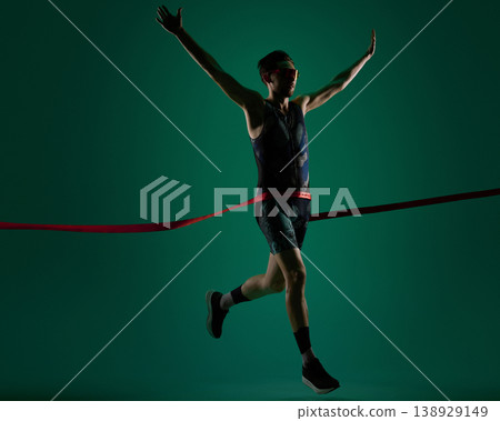 Male athlete crossing finish line with raised arms on dark green background. 138929149