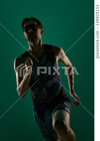 Close up of male athlete running toward camera in dark green studio lighting. 138929153