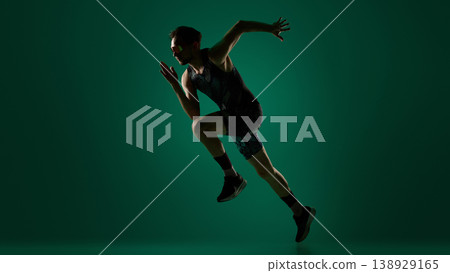 Male athlete performing dynamic sideways jump with extended limbs on dark green background. Male athlete performing dynamic sideways jump with extended limbs on dark green background. 138929165