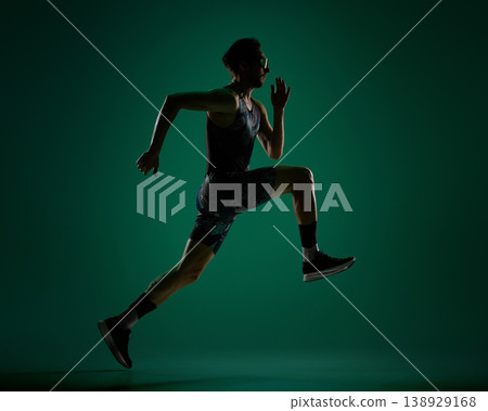 Male athlete jumping forward mid air in dynamic sprint motion on dark green background. 138929168