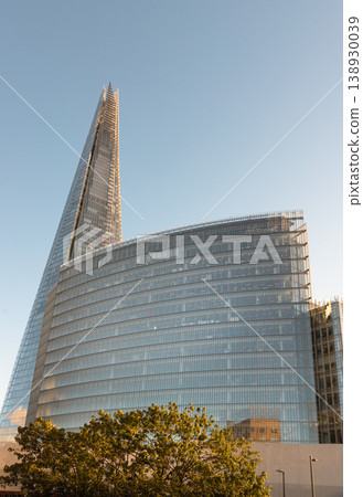 The Shard pierces the horizon as a soaring needle of glass, standing as a dominant silhouette against the sky. Beside it, the gracefully curved News Building reflects the sun golden glow, its rhythmic 138930039