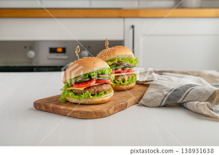 Two beef burgers with tomato, cheese, and lettuce on a cutting board on a light table with napkin Two beef burgers with tomato, cheese, and lettuce on a cutting board on a light table with napkin 138931638
