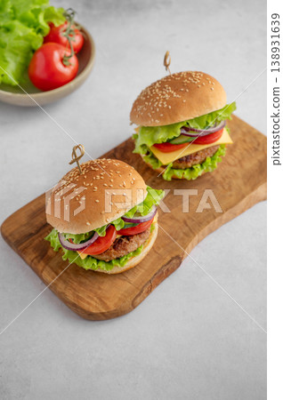 Two beef burgers with tomato, cheese, and lettuce on a cutting board on a light textured background Two beef burgers with tomato, cheese, and lettuce on a cutting board on a light textured background 138931639