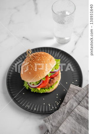 Beef burger with tomato, cheese, and lettuce on a plate on a marble background with glass of water Beef burger with tomato, cheese, and lettuce on a plate on a marble background with glass of water 138931640