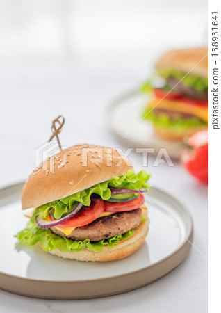 Two Beef burgers with tomato, cheese, and lettuce on a plate on a white table near window. Two Beef burgers with tomato, cheese, and lettuce on a plate on a white table near window. 138931641