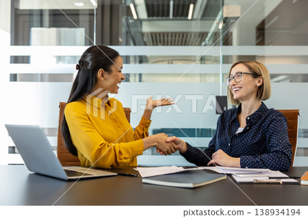 Two businesswomen are smiling and shaking hands across an office desk with a laptop and documents, symbolizing a successful partnership, collaboration, and agreement Two businesswomen are smiling and shaking hands across an office desk with a laptop and documents, symbolizing a successful partnership, collaboration, and agreement 138934194