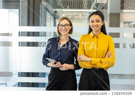Two smiling professional business women, one holding a tablet and another with crossed arms, standing confidently in a bright contemporary office, illustrating professionalism, teamwork, and success Two smiling professional business women, one holding a tablet and another with crossed arms, standing confidently in a bright contemporary office, illustrating professionalism, teamwork, and success 138934209