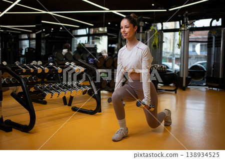 Young adult woman doing dumbbell lunges in modern gym 138934525