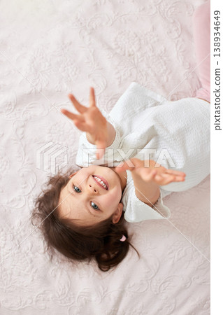 Little girl smiling and reaching hands up while lying on a cozy bed, embodying childhood joy and innocence 138934649