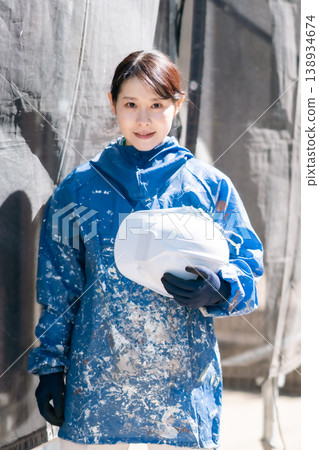 A smiling female painter holding a helmet. Architectural painting, maintenance, skilled worker, female on site, outdoors. A smiling female painter holding a helmet. Architectural painting, maintenance, skilled worker, female on site, outdoors. 138934674