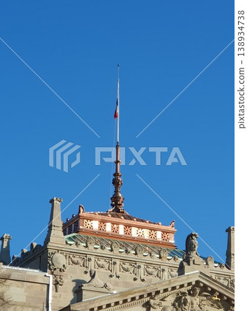 Historic ornate building rooftop featuring a copper spire and a small flag pole set against a vivid clear blue sky, showcasing architectural details, sculptures, and classical decorative elements. Historic ornate building rooftop featuring a copper spire and a small flag pole set against a vivid clear blue sky, showcasing architectural details, sculptures, and classical decorative elements. 138934738