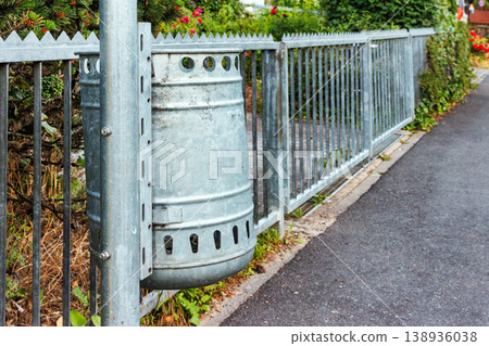 Galvanized metal trash bin attached to a pole with a metal fence next to a sidewalk. Urban street furniture for public waste disposal and cleanliness concepts. Galvanized metal trash bin attached to a pole with a metal fence next to a sidewalk. Urban street furniture for public waste disposal and cleanliness concepts. 138936038