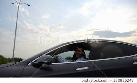 Young businessman driving on electric car in circular path on asphalt road after rain. Happy manager showing grimaces and fooling around while riding on modern auto. Concept of green innovation Young businessman driving on electric car in circular path on asphalt road after rain. Happy manager showing grimaces and fooling around while riding on modern auto. Concept of green innovation 138936242
