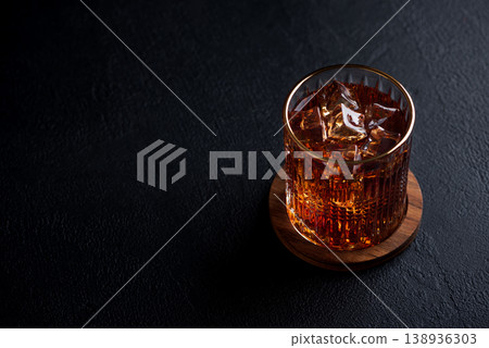Whiskey Glass with Ice on Wooden Coaster against Dark Concrete Background 138936303