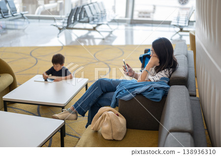 Woman Using Smartphone While Child Engages with Tablet in Modern Airport Lounge Woman Using Smartphone While Child Engages with Tablet in Modern Airport Lounge 138936310