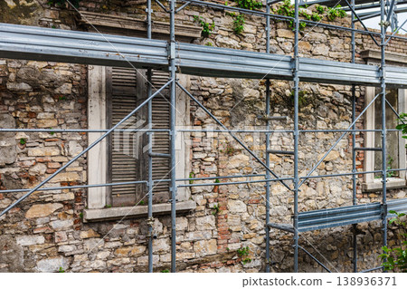 Old stone wall with wooden window shutters covered by metal scaffolding during building renovation in Italy Old stone wall with wooden window shutters covered by metal scaffolding during building renovation in Italy 138936371