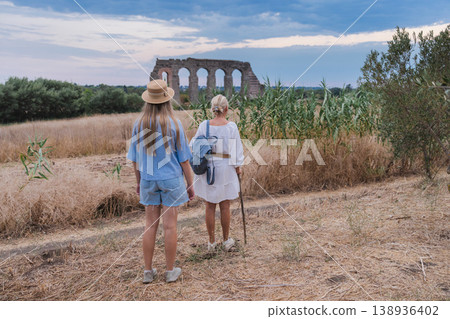 Mother and teenage daughter walking toward ancient Roman aqueduct in Parco degli Acquedotti Rome at sunset through dry grass and natural landscape Mother and teenage daughter walking toward ancient Roman aqueduct in Parco degli Acquedotti Rome at sunset through dry grass and natural landscape 138936402