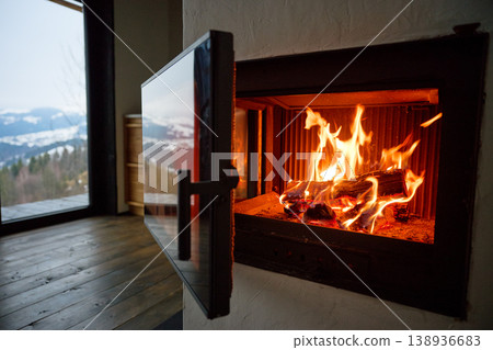 Fire burns in modern fireplace inside cozy home. Close up of glowing logs and flames, glass door open, wooden floor and mountain view through window, warmth and comfort concept Fire burns in modern fireplace inside cozy home. Close up of glowing logs and flames, glass door open, wooden floor and mountain view through window, warmth and comfort concept 138936683