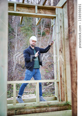 Construction worker measuring wall height with tape inside wooden frame house, showing OK gesture while confirming correct dimensions during building process Construction worker measuring wall height with tape inside wooden frame house, showing OK gesture while confirming correct dimensions during building process 138936691