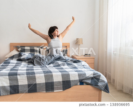 An adult woman feeling refreshed and well-rested, stretching her arms and waking up to natural light in a cozy bedroom. Authentic lifestyle An adult woman feeling refreshed and well-rested, stretching her arms and waking up to natural light in a cozy bedroom. Authentic lifestyle 138936695