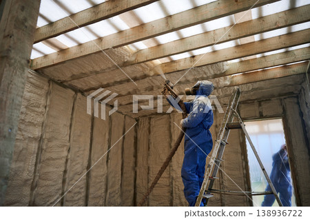 Installer covering overhead framing with spray foam insulation in house under construction. Ladder access, protective suit and energy saving building envelope concept Installer covering overhead framing with spray foam insulation in house under construction. Ladder access, protective suit and energy saving building envelope concept 138936722