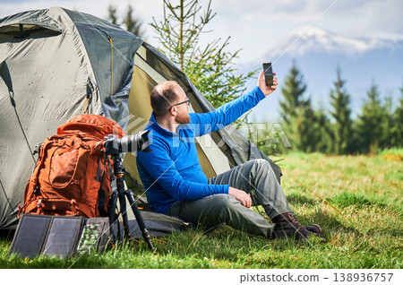 Man takes selfie with smartphone outside tent in mountain campsite with camera gear, backpack, and portable solar panel. Useful for outdoor travel, remote work, and adventure marketing Man takes selfie with smartphone outside tent in mountain campsite with camera gear, backpack, and portable solar panel. Useful for outdoor travel, remote work, and adventure marketing 138936757