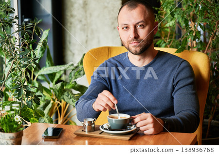 Man stirring coffee and looking at camera in cozy cafe, calm lifestyle scene, relaxed break, modern urban living, everyday routine, quiet moment with drink Man stirring coffee and looking at camera in cozy cafe, calm lifestyle scene, relaxed break, modern urban living, everyday routine, quiet moment with drink 138936768