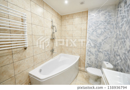Modern bathroom interior with corner bathtub, toilet and sink. Bright washroom with beige wall tiles, textured accent wall, heated towel rail and clean minimalist design Modern bathroom interior with corner bathtub, toilet and sink. Bright washroom with beige wall tiles, textured accent wall, heated towel rail and clean minimalist design 138936794