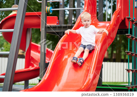 Child sliding down playground slide, holding sides , summer outfit, bright red slide and play structure, outdoor park, active play, childhood fun Child sliding down playground slide, holding sides , summer outfit, bright red slide and play structure, outdoor park, active play, childhood fun 138936819