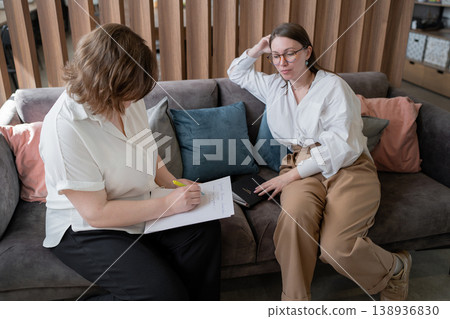 Women sitting on sofa reviewing papers and collaborating in cozy workplace lounge. Collaboration, reviewing, papers, lounge, women, business. 138936830