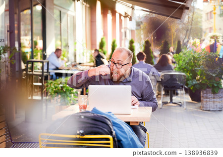 Man eating meal at outdoor cafe while working on laptop, sitting among other visitors, focused on screen, combining lunch and remote work in busy urban environment Man eating meal at outdoor cafe while working on laptop, sitting among other visitors, focused on screen, combining lunch and remote work in busy urban environment 138936839